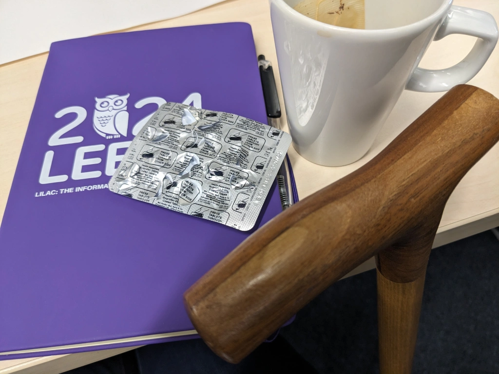Three items on a desk: a purple notebook with the LILAC 2024 logo on it, an empty cop of tea, and a packet of ibuprofen. Leaning against the desk is a wooden walking stick.